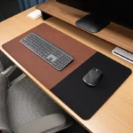 Reddish brown executive leather desk mat with black fabric mouse area placed on a modern office desk with keyboard and monitor stand
