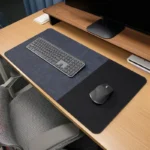 Blue gray leather desk mat with black fabric mouse area shown on a wood office desk with keyboard, mouse, and monitor riser