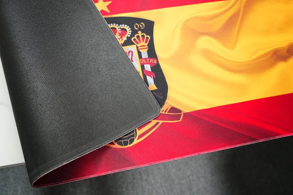 Spain football desk mat lifted to show black anti-slip rubber backing, flexible material, and stitched edge construction.